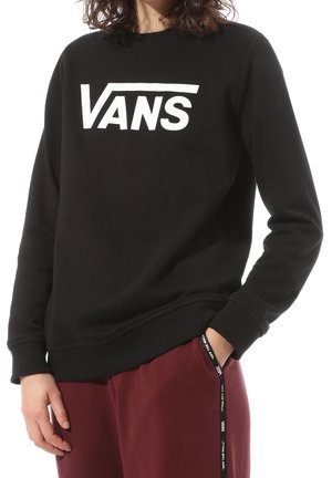 Persona con sudadera negra Vans con logo blanco y pantalones granate, una mano en el bolsillo, sobre fondo blanco.