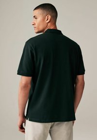 Camisa polo verde oscuro de manga corta, con un cuello plano y aberturas laterales para facilitar el movimiento. Textura de tela suave.