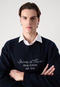 Tengerész pulóver puha anyagból, kerek nyakkal, hímzett "Sports d'Hiver FROM FUTURE EST. 2018" felirattal és bordás mandzsettával.