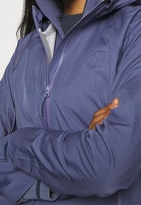 Fioletowa wodoodporna kurtka z zamkiem błyskawicznym z przodu, wysokim kołnierzem i polarowym wykończeniem. Posiada dopasowane mankiety oraz detal z logo na piersi.