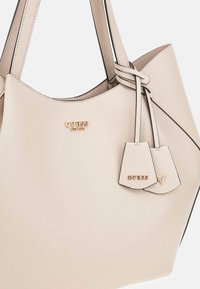 Borsa a tracolla in finta pelle beige con logo dorato, design strutturato, texture sottili e due etichette decorative attaccate con cinturini sottili.