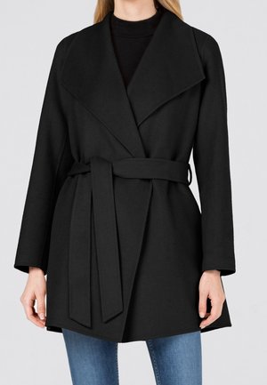 Femme portant un manteau noir ceinturé à larges revers sur un jean bleu, debout devant un fond gris clair.