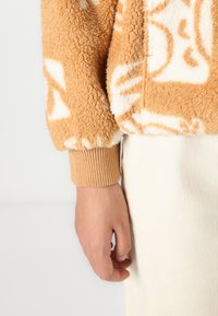 Fleece-pullover i varm ferskenfarge med hvite abstrakte mønstre. Har ribbestrikkede mansjetter og en myk, strukturert overflate. Nærbilde av ermet og hånden.