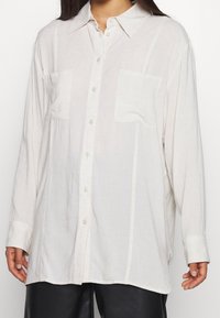 Camicia ampia a maniche lunghe in tessuto beige chiaro, con chiusura a bottoni sul davanti, due tasche sul petto e sottili righe verticali.