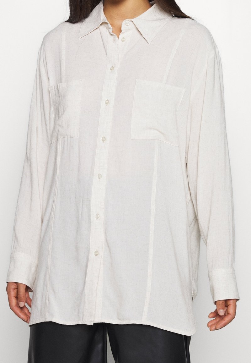 Camicia ampia a maniche lunghe in tessuto beige chiaro, con chiusura a bottoni sul davanti, due tasche sul petto e sottili righe verticali.