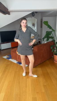 Young woman in black-and-white striped dress and white flats standing in a modern living room with wooden floor and brown couch.