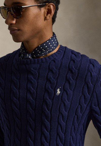 Muškarac koji nosi sunčane naočale u stilu oklopa kornjače, plavi kabelski džemper s malim logotipom igrača pola i plavu točkastu maramu na vratu.