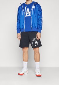 Sininen vetoketjullinen takki hupulla, valkoista tekstiä; sininen t-paita, jossa LA-logo; mustat shortsit, joissa valkoinen LA-printti; valkoiset sneakersit punaisilla yksityiskohdilla.