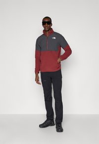 Rød og grå lynlås fleece-pullover med høj krave, parret med sorte bukser og sorte sko, sat mod en ensfarvet baggrund.