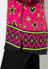 Heldere roze stof met gele en zwarte sterren en harten, geometrisch randpatroon, gedragen over zwarte broek met een zijsplit.
