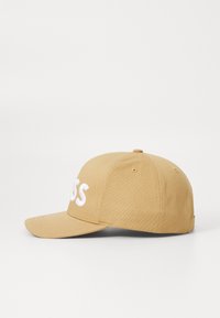 Beige színű baseball sapka ívelt napellenzővel, elöl fehér hímzett szöveggel. Az anyag sima felületű és strukturált kialakítású.