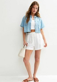 Camisa de mezclilla azul claro, sin mangas blanca, pantalones cortos blancos sueltos, sandalias marrones y bolso pequeño beige, con texturas suaves y diseño casual.
