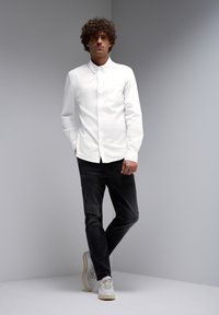 Camisa blanca de botones con un bolsillo en el pecho, combinada con jeans de mezclilla oscuros y zapatillas blancas. El modelo se encuentra de pie frente a un fondo neutral.