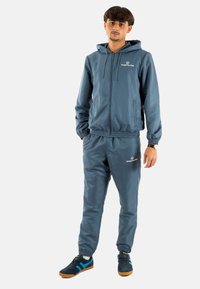 Chándal ligero azul con puños y cinturilla el�ásticos, que presenta una chaqueta con cremallera, capucha y detalle de logo. Combinado con zapatillas deportivas.