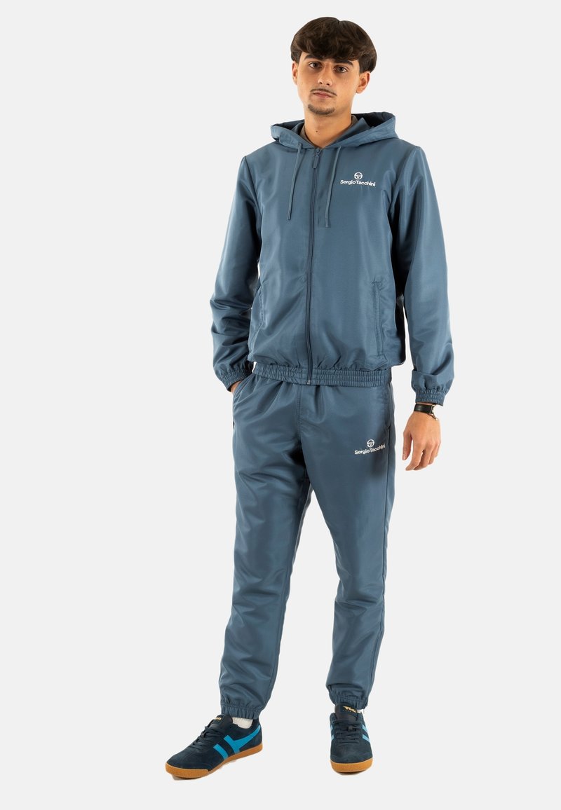 Chándal ligero azul con puños y cinturilla elásticos, que presenta una chaqueta con cremallera, capucha y detalle de logo. Combinado con zapatillas deportivas.