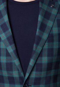 Zielono-granatowa marynarka w kratę z czerwonymi akcentami, z teksturowanej tkaniny oraz z pojedynczym zapięciem na guzik, nałożona na ciemnogranatowy t-shirt.