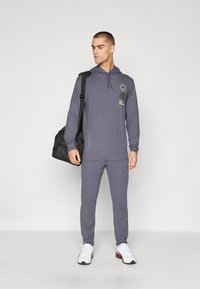 Grå hettegenser med lange ermer og frontlogo-design, sammen med matchende grå joggebukser. Modellen bærer en svart weekendbag og har på seg hvite sneakers.
