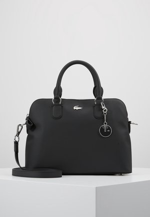 Sac à main en cuir noir avec deux anses, bandoulière amovible, logo Lacoste argenté, et porte-clés rond noir, présenté sur un piédestal blanc.