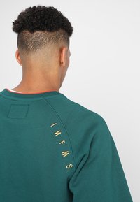 Zielony bawełniany sweatshirt z okrągłym dekoltem. Posiada złoty haftowany tekst na ramieniu oraz teksturowane wykończenie materiału.