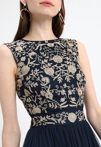 Mörkblå klänning med intrikata gyllene blommönster på midjan och en plisserad kjol. Ärmlös design med en rund halsringning.