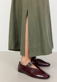 Gonna verde oliva, lunghezza alla caviglia, con spacco laterale. La persona indossa scarpe in pelle bordeaux con dettaglio della cintura e fibbia. Tessuto liscio, stile attillato.