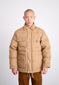 Braune Steppjacke mit einem Cordkragen, zwei Vorderaschen und einem Logo-Patch. Der Stoff ist strukturiert und gesteppte für Wärme.