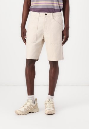 Beżowe bawełniane szorty o luźnym kroju, z przednimi kieszeniami i zapięciem na guzik, zestawione z beżowymi trampkami i szarymi skarpetkami.