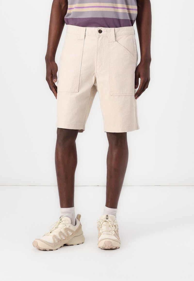 Shorts en coton beige avec une coupe décontractée, dotés de poches avant et d'une fermeture à bouton, associés à des baskets beiges et des chaussettes grises.