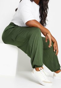 Personne portant un pantalon cargo vert olive, des baskets blanches et une chemise blanche à manches courtes, assise sur un bloc blanc avec un bras reposant sur son genou.