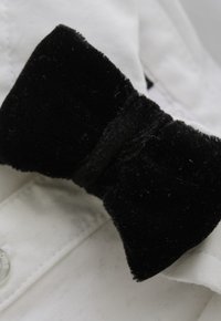 Zwarte fluwelen vlinderdas met een zachte textuur, met een centrale knoop, rustend tegen een wit overhemd. De gladde stof contrasteert met de textuur van de vlinderdas.