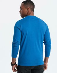 Man som har på sig en ljusblå långärmad skjorta och svarta jeans, vänd bort, med en svart klocka på vänster handled mot en enfärgad bakgrund.