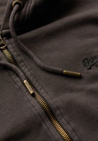 Sudadera con cremallera de color gris oscuro, con un zipper en bronce y extremos de cordón, con el logo "Superdry" sutilmente bordado en la tela.