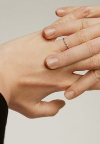 Delicado anillo de plata con un diseño ondulado, adornado con pequeñas piedras preciosas transparentes, que se lleva en una mano con esmalte de uñas natural.