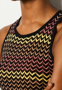 Stickad linne med ett zickzackmönster i svarta, gula, röda och bruna nyanser. Bred halsringning och textil med en strukturerad yta och intrikata detaljer.