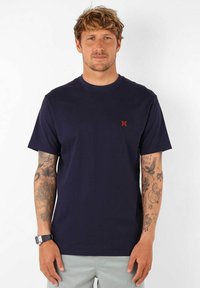 T-shirt en coton bleu marine avec un col rond, des manches courtes et un petit logo rouge sur la poitrine gauche. Texture lisse et design classique.