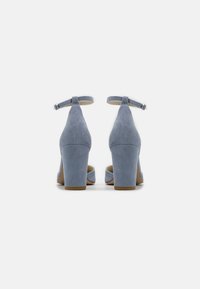 Zapatos de tacón alto de gamuza azul con correas en el tobillo, que cuentan con un tacón grueso y un diseño de punta abierta. Superficie texturizada y suela en un tono marrón claro.