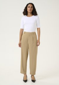 Beige hoog-getailleerde broeken met plooien, gecombineerd met een fitted witte top. Model draagt zwarte pumps met een spitse neus. Eenvoudig, recht model.