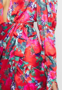 Robe florale rouge avec un décolleté en V profond, de longues manches bouffantes et une ceinture à la taille. Le tissu est en satin avec des fleurs éclatantes de rose, vert et bleu.