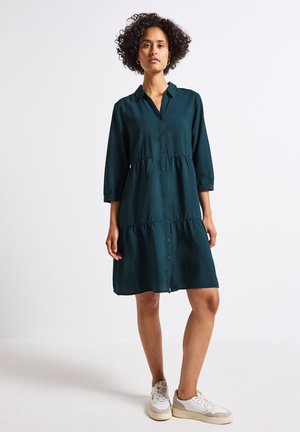 Grünes, knopfbares Kleid mit gestuften Design, Dreiviertelärmeln und lockerem Schnitt; dezente Textur. Getragen mit weißen Turnschuhen.
