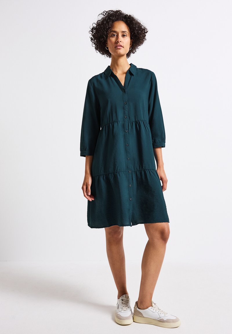 Robe verte à boutons avec un design en étages, manches trois quarts et coupe décontractée ; texture subtile. Portée avec des baskets blanches.