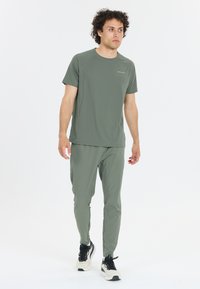 Man med lockigt hår som går, iklädd en olivgrön sporttröja och byxor med vita och svarta sneakers mot en vit bakgrund.