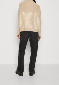 Beżowy sweter z prążkowanym wzorem w białe poziome paski, w połączeniu z czarnymi prostymi spodniami i czarnymi botkami, widok z tyłu.