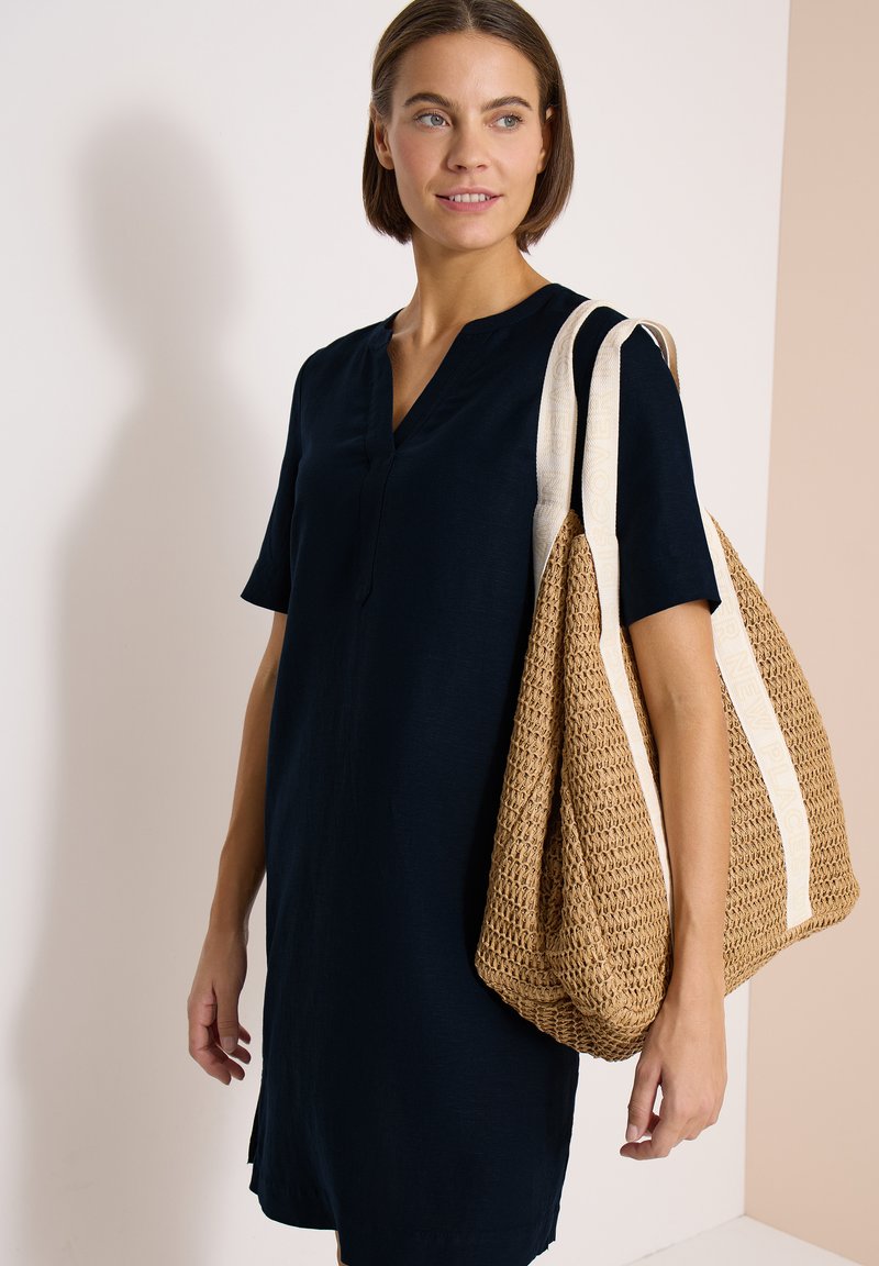Vrouw in zwarte jurk draagt een grote geweven beige draagtas met brede witte riemen over haar schouder, kijkt opzij met een lichte glimlach.