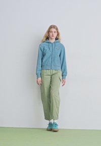 Sudadera de cremallera azul claro con una cintura ajustada, combinada con pantalones cargo verdes holgados. El modelo lleva zapatos teal y calcetines blancos sobre un suelo verde.