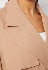 Beżowy trencz z dużym klapowym kołnierzem, wykonany z gładkiej tkaniny, z widocznymi detalami przeszyć i luźnym krojem.