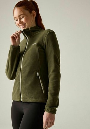 Joven sonriente cerrando una chaqueta polar verde oliva con bolsillos con cremallera, vistiendo pantalones negros, de pie frente a un fondo liso.