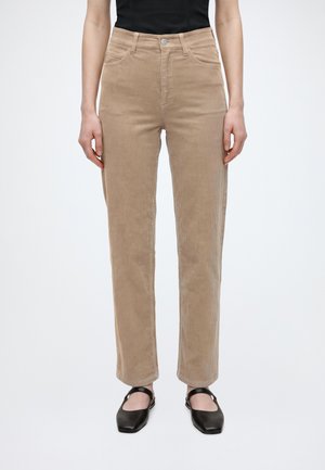 Persoon die beige ribfluwelen broek, zwart topje en zwarte Mary Jane schoenen draagt, staand tegen een effen lichte achtergrond.
