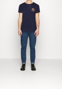 Camiseta de algodón azul marino con cuello redondo, que presenta un logo bordado en el pecho. Combinada con unos vaqueros ajustados de mezclilla azul y botas negras.