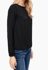 Svart långärmad blus med rund halsringning, med en subtil textur och knappstängning vid halsen och ärmsluten. Kombineras med blå jeans.