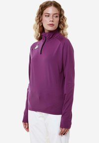 Donna con capelli biondi mossi che indossa un top atletico viola a maniche lunghe con una cerniera nera e pantaloni bianchi, in piedi su uno sfondo uniforme.